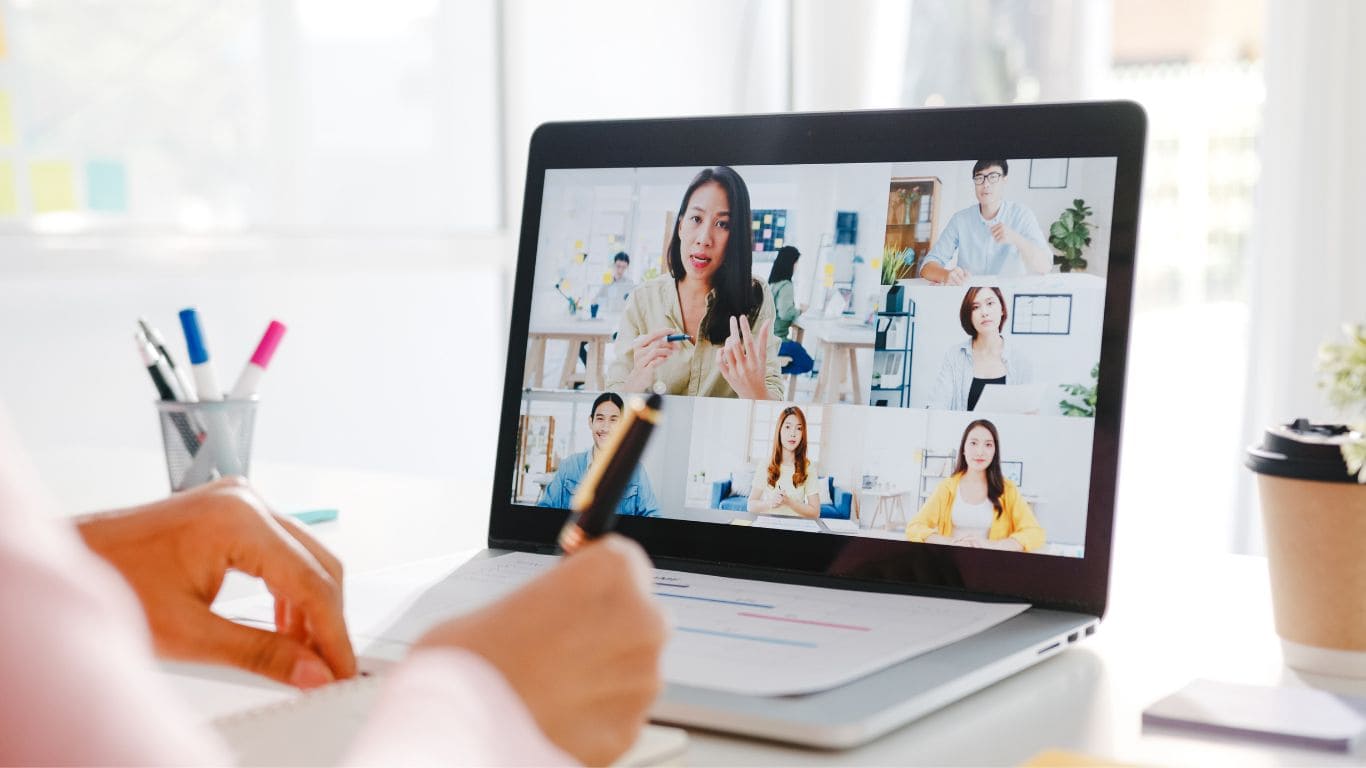 Imagem de uma mulher participando de uma vídeo conferência com outras mulheres, ela utiliza um notebook e faz anotações